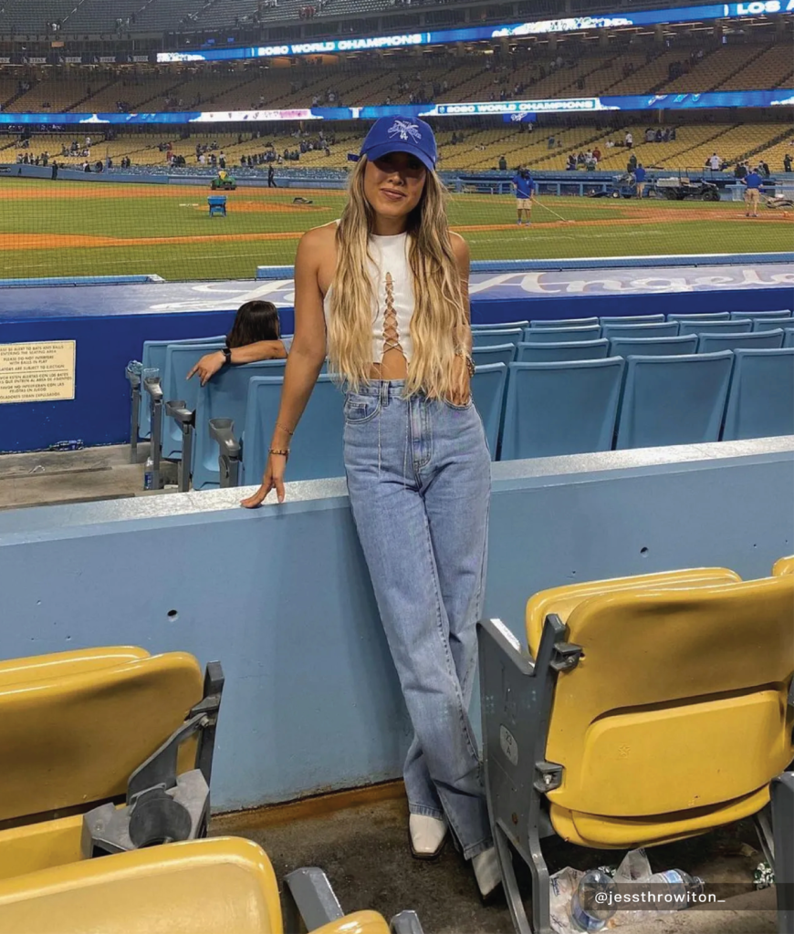 woman wearing white tank top, relaxed blue jeans, ankle boots, and baseball cap at stadium
