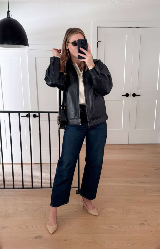 Woman wearing a cream cashmere cardigan under a black leather jacket, paired with dark barrel-leg denim jeans and nude kitten heels, standing on a city sidewalk.