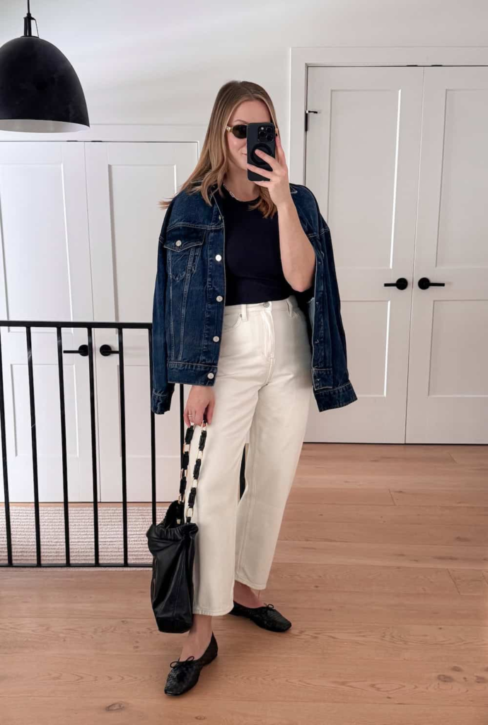 Woman wearing ecru jeans with a black tee, an oversized dark denim jacket, and black woven leather flats, styled with a matching handbag.