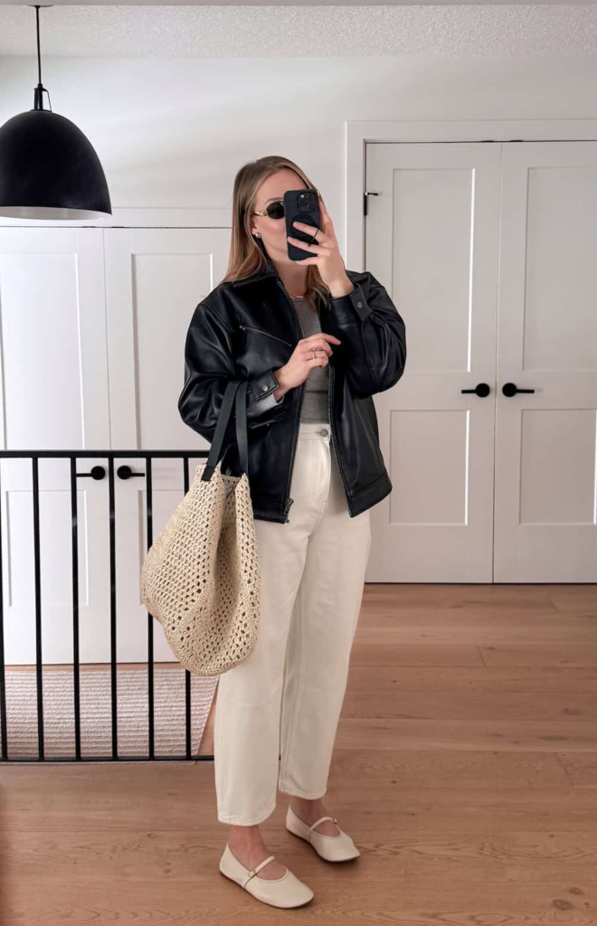 Woman wearing a black leather bomber jacket with light-wash jeans, a neutral knit top, white Mary Jane flats, and carrying a woven tote bag.