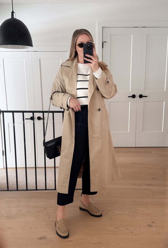 Woman wearing a black and white striped knit sweater with tailored black trousers, a beige trench coat, and suede loafers.