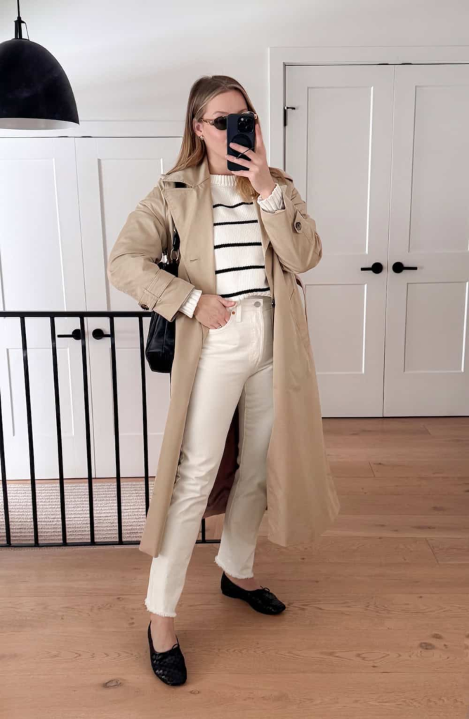 Woman wearing a striped sweater with white jeans, a beige trench coat, and black ballet flats, styled for a casual spring day.