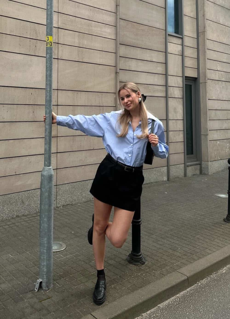 Woman wearing a mini skirt with a light blue button-up shirt, styled with loafers and socks for a casual spring outfit.