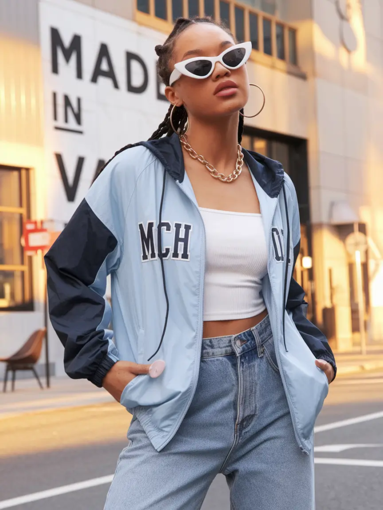 Lightweight blue and navy windbreaker layered over a white ribbed tank with light denim jeans and statement accessories