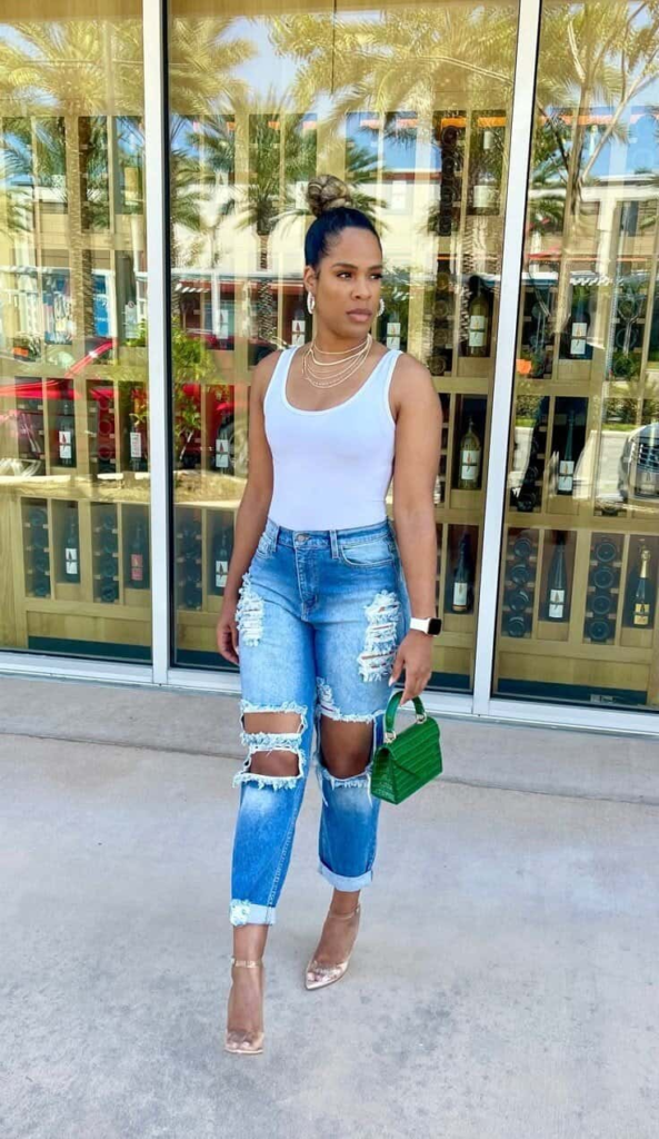 Woman wearing a white tank top tucked into heavily distressed light-wash jeans, accessorized with a green mini handbag, layered gold necklaces, and clear heels, standing on a sunny boardwalk.