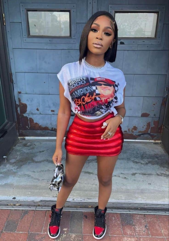 Woman wearing an oversized graphic tee tucked into a red mini skirt, paired with red and black sneakers and a sleek bob hairstyle, standing on a sunlit urban street.