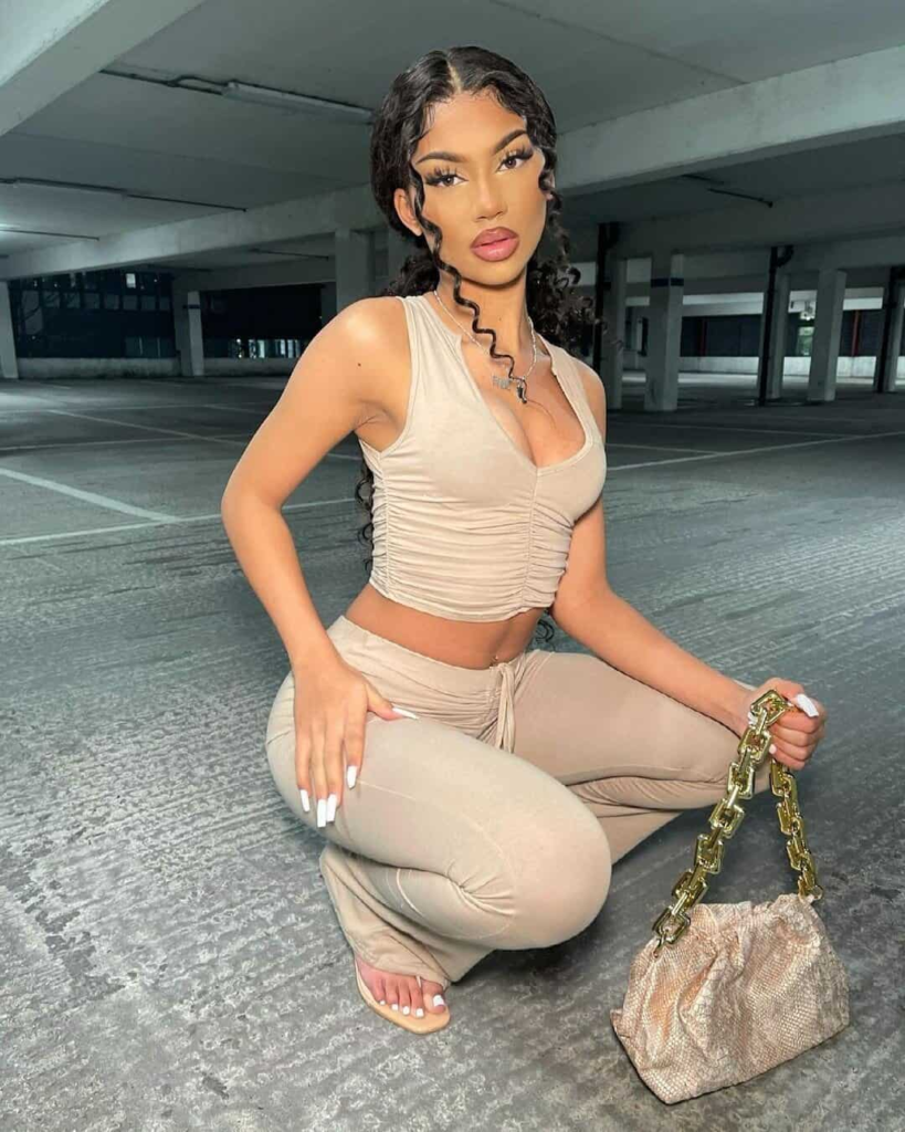 Woman wearing a nude ruched crop top with high-waisted nude pants, accessorized with a gold chain handbag and delicate bangles, standing on a sunlit resort terrace.