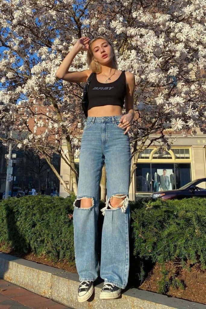 Woman wearing a black crop top with bold text paired with light-wash high-waisted ripped wide-leg jeans and classic sneakers, standing on a sunlit urban street.