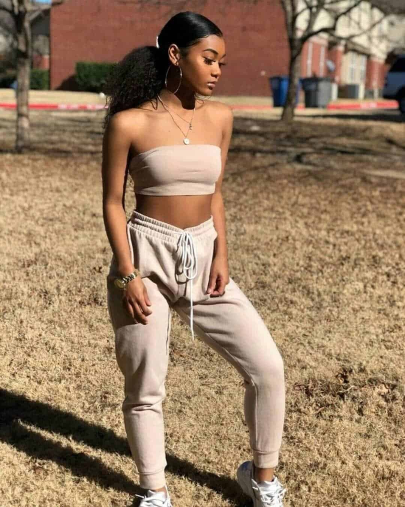 Woman wearing a neutral strapless crop top paired with high-waisted matching joggers, accessorized with hoop earrings and layered necklaces, standing in a sunlit urban plaza.