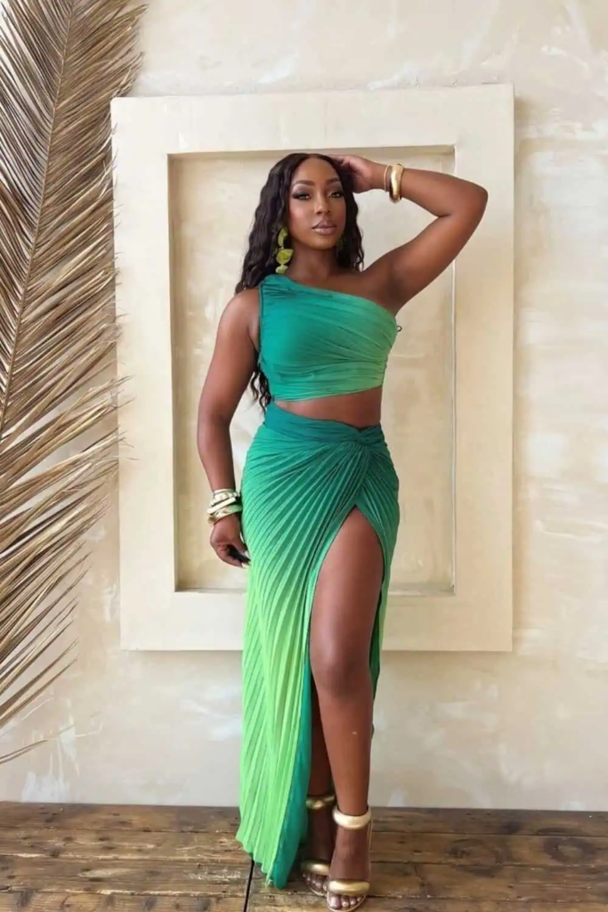 Woman wearing a one-shoulder green crop top and high-slit ruched maxi skirt in ombré emerald to lime, accessorized with gold bangles and strappy metallic heels, standing on a sunlit tropical terrace.