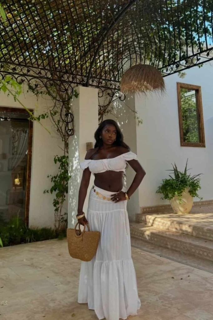 Woman wearing an off-shoulder white crop top and flowing white maxi skirt with a gold coin belt, accessorized with a woven beach bag and gold bangles, standing on a sunlit tropical terrace.