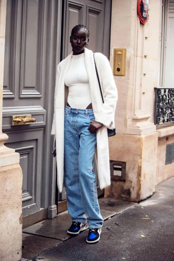 Woman wearing a long white coat over a white bodysuit and relaxed-fit jeans, accessorized with sneakers and delicate gold jewelry, standing on a sunlit urban street.