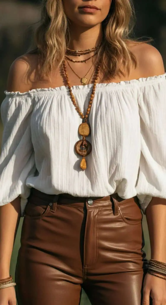 Woman wearing an off-shoulder embroidered peasant blouse tucked into fitted brown leather pants, accessorized with layered necklaces, statement rings, wedge sandals, and a tooled leather bag, standing on a sunlit patio.