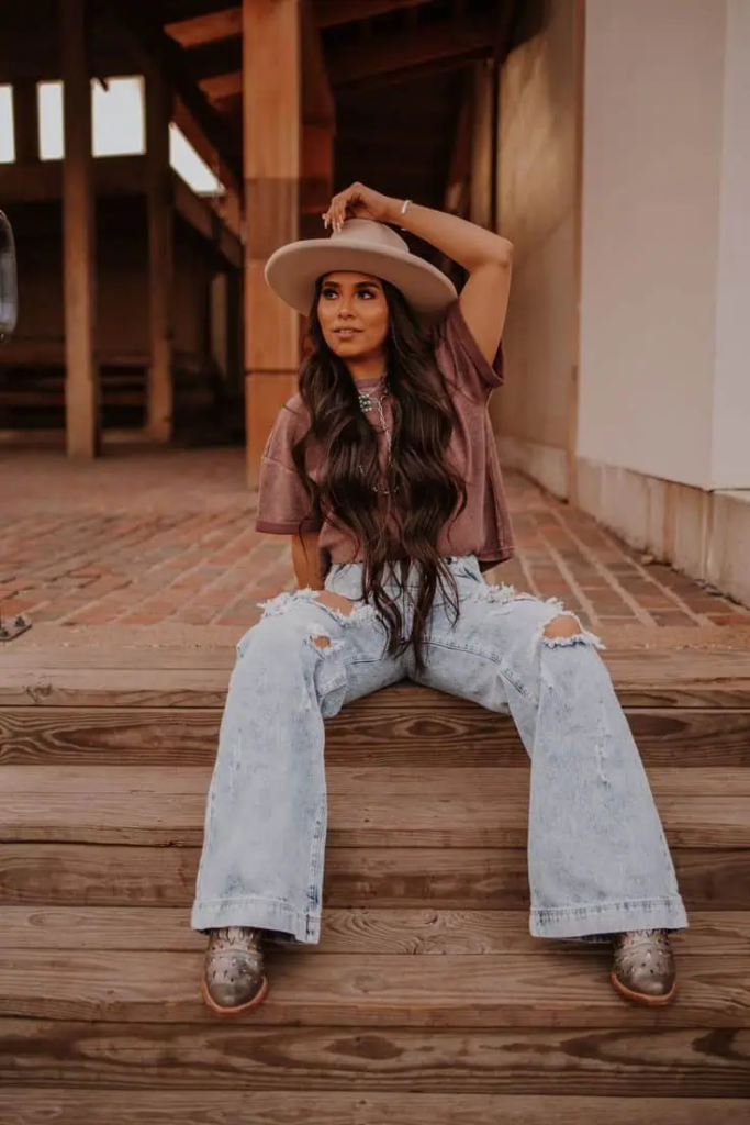 Woman wearing mauve cropped top, distressed wide-leg jeans, metallic ankle boots, and wide-brim hat, styled for a Western fall look.