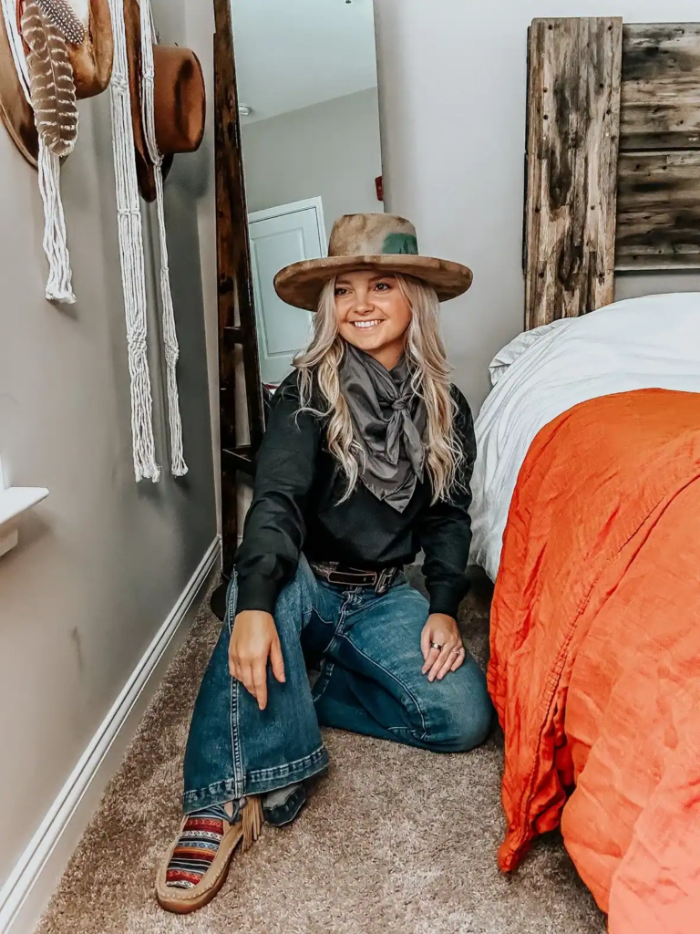 Woman in black long-sleeve top, blue jeans, gray silk neckerchief, earth-toned wide-brim hat, and patterned moccasins in Western fall setting.