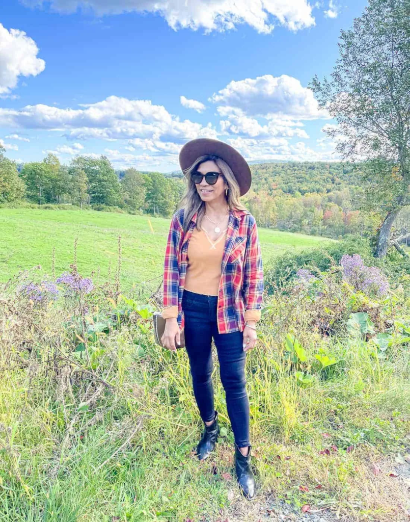 Western-inspired outfit with plaid shirt over orange top, dark jeans, black ankle boots, wide-brim hat, and sunglasses outdoors.