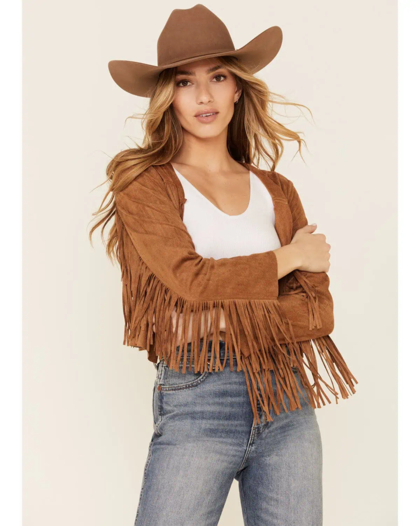 Woman wearing caramel suede fringe jacket, white tank, blue jeans, and wide-brim cowboy hat in fall setting.