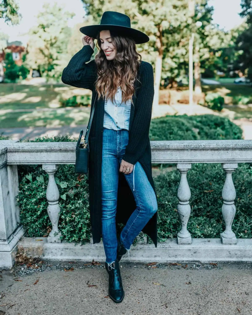 Woman in black long cardigan over denim shirt and jeans, black ankle boots, and wide-brim hat outdoors.