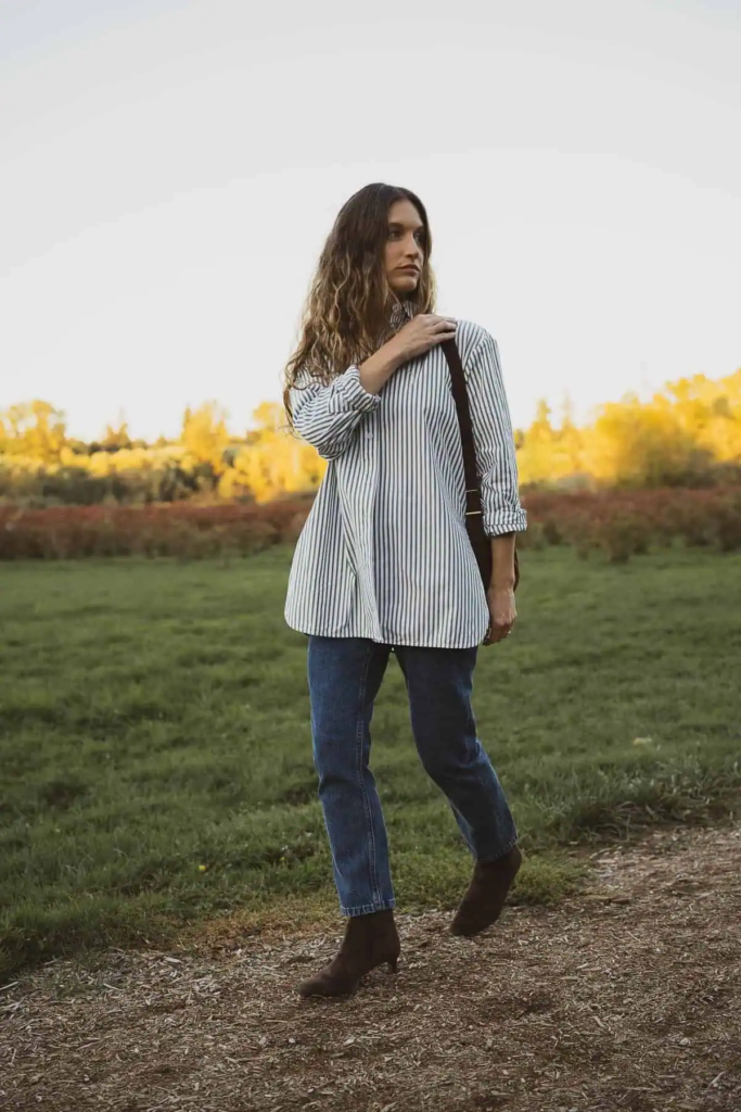 Woman wearing oversized striped shirt with straight-leg denim jeans, brown ankle boots, and wide-brim hat outdoors in autumn.