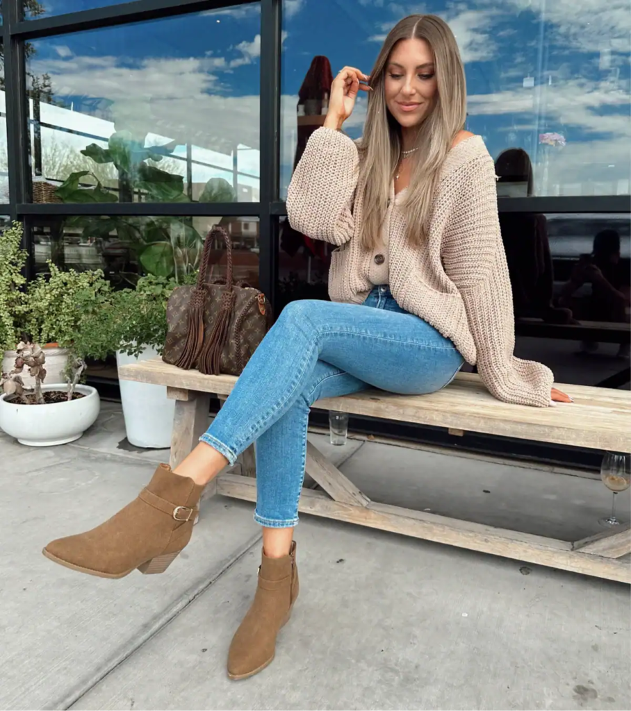 Woman wearing oversized chunky knit cardigan, blue jeans, suede ankle boots, and fringe handbag in autumn setting.
