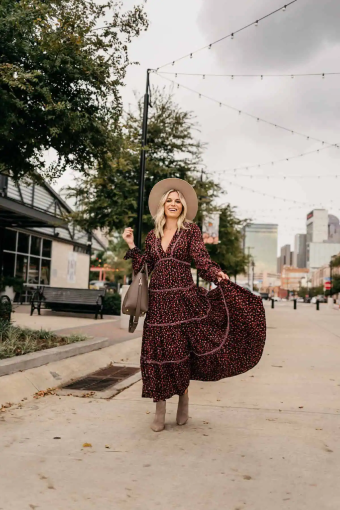 Woman wearing burgundy floral long-sleeved maxi dress, suede ankle boots, and wide-brim felt hat in fall outdoors.