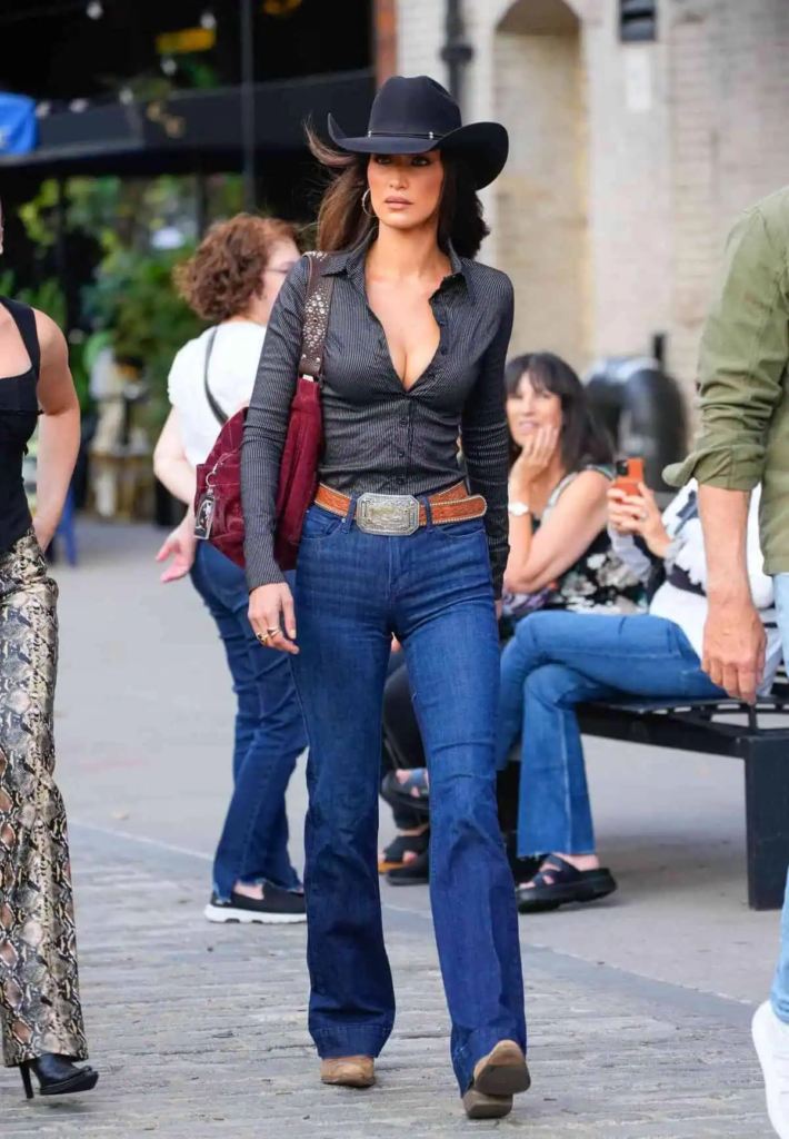 Woman wearing fitted pinstripe shirt, high-waisted flared jeans, burgundy bag, and wide-brim black hat in urban Western fall outfit.