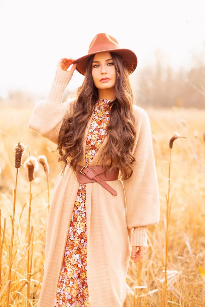 Woman wearing cream cardigan layered over floral dress with rust wide-brim hat and brown leather boots in autumn field.