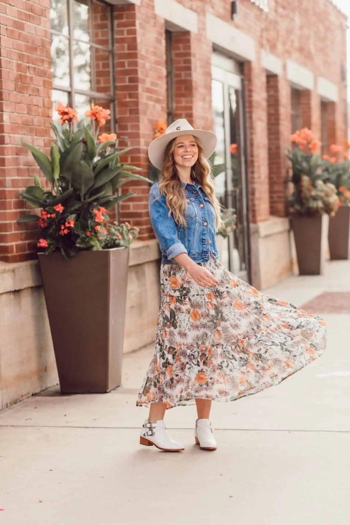 Woman wearing light denim jacket with floral midi skirt, white ankle boots, and beige wide-brim hat outdoors in fall.