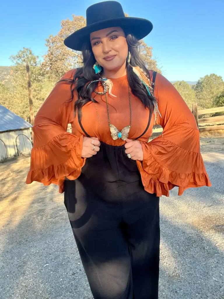 Woman wearing burnt orange bell-sleeve top, dark high-waisted pants, turquoise jewelry, and black wide-brim hat in Western fall outfit.