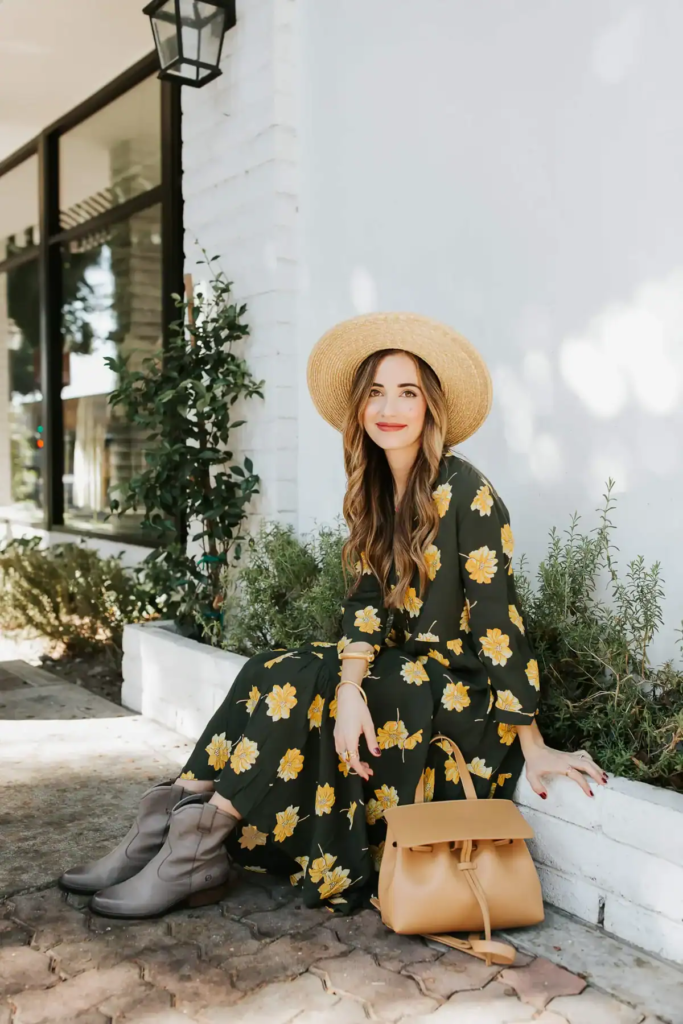 Woman wearing green floral maxi dress with Western ankle boots, straw wide-brim hat, and leather handbag in fall setting.