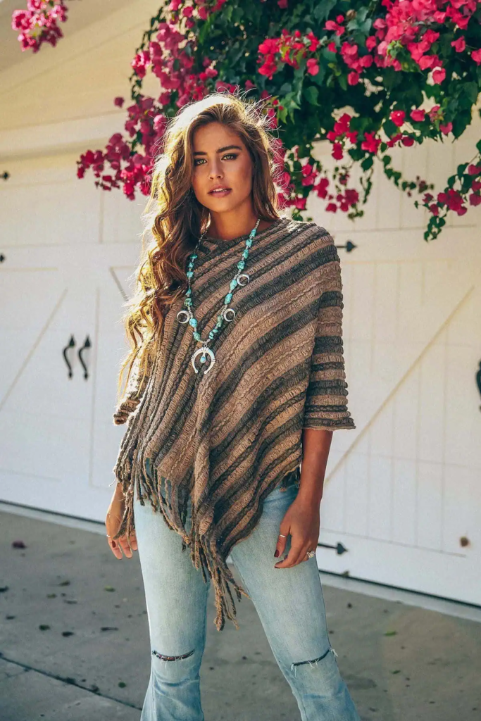 Woman wearing a warm-toned fringed poncho over light-wash jeans, accented with a turquoise necklace and taupe fringe bag.