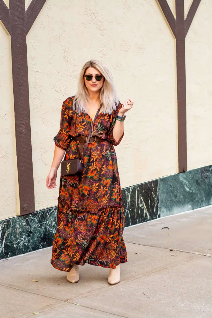 Woman in a burnt orange and deep purple floral maxi dress, cinched with a brown leather belt, paired with neutral ankle boots.