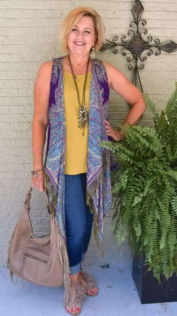 Woman wearing a purple paisley vest over a mustard top with blue jeans, accessorized with layered necklaces and bracelets.