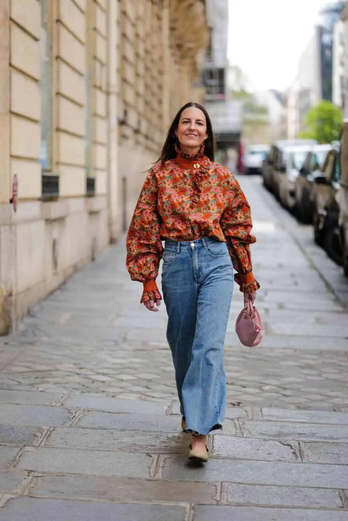 Woman wearing a warm-toned floral blouse tucked into wide-leg blue jeans, carrying a small handbag.
