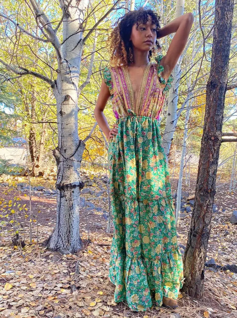 Woman in a green and pink floral maxi dress standing among autumn foliage, wearing simple shoes and minimal jewelry.