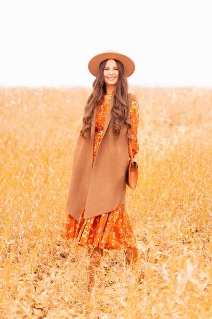 Woman wearing an orange floral maxi dress layered with a camel sleeveless coat and a wide-brim hat, standing in a golden wheat field.
