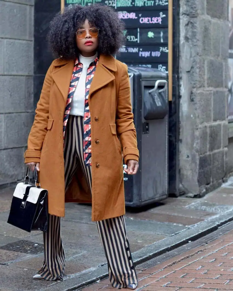 Woman wearing a camel coat over a striped pant and printed shirt outfit, accessorized with a white turtleneck and structured handbag.