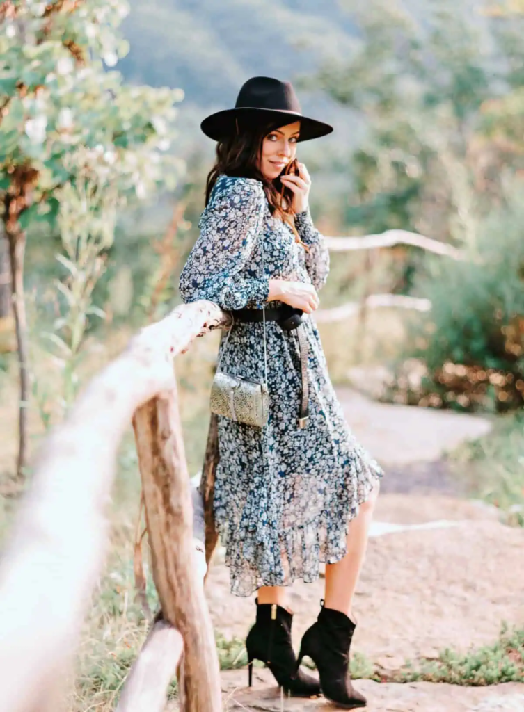 Woman wearing a muted floral dress with a wide-brim black hat, styled with black ankle boots and a metallic crossbody bag.